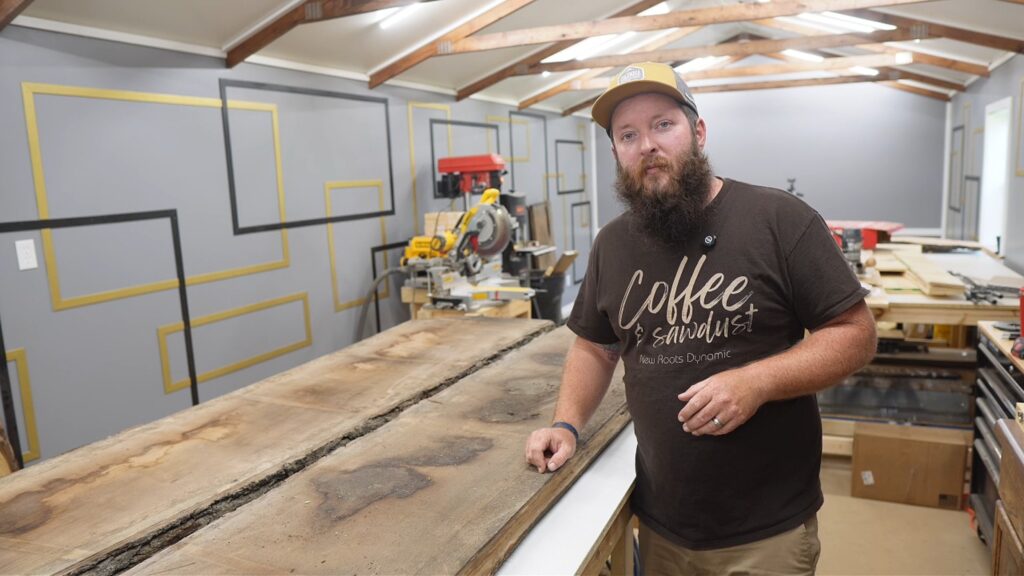 Curtis in his workshop wearing a Coffee and sawdust New Roots Dynamic shirt, hand resting on walnut slabs, wide shop shot with tools behind him.