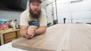 Close-up of Curtis leaning on a walnut slab in the workshop and talking to the camera.